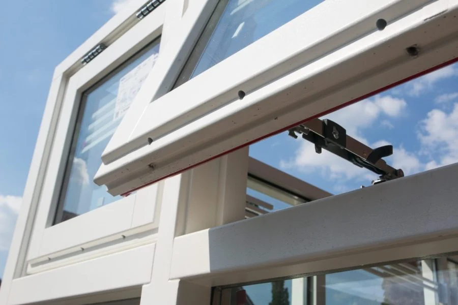 Close-up van een openstaand uitzetraam met zwart raamuitzetter in wit kozijn, gemonteerd in een glazen serreconstructie – Toelevering Online